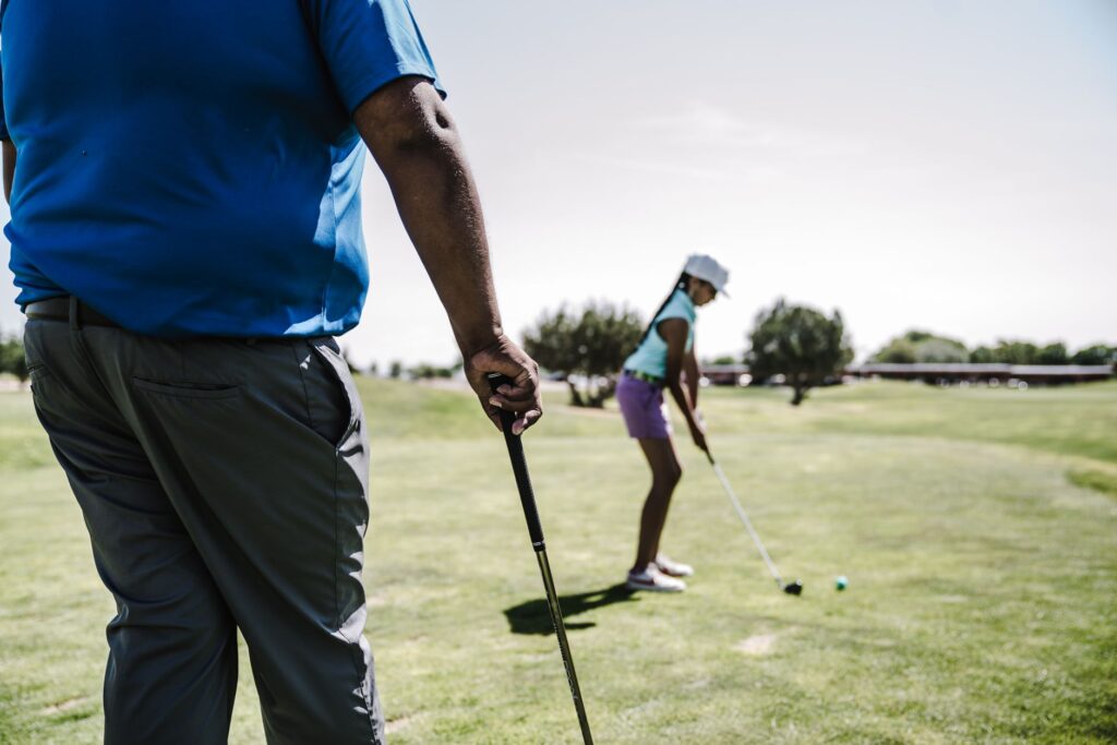 A child holding a golf club with a parent, both ready to tee off on a sunny day. — family golf Dungeness